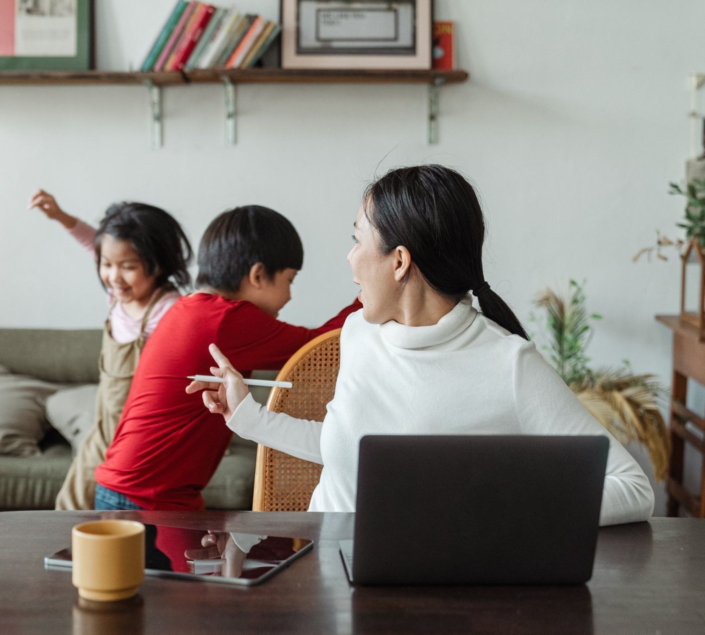 A woman is sitting at a table with a laptop and two children playing in the background.