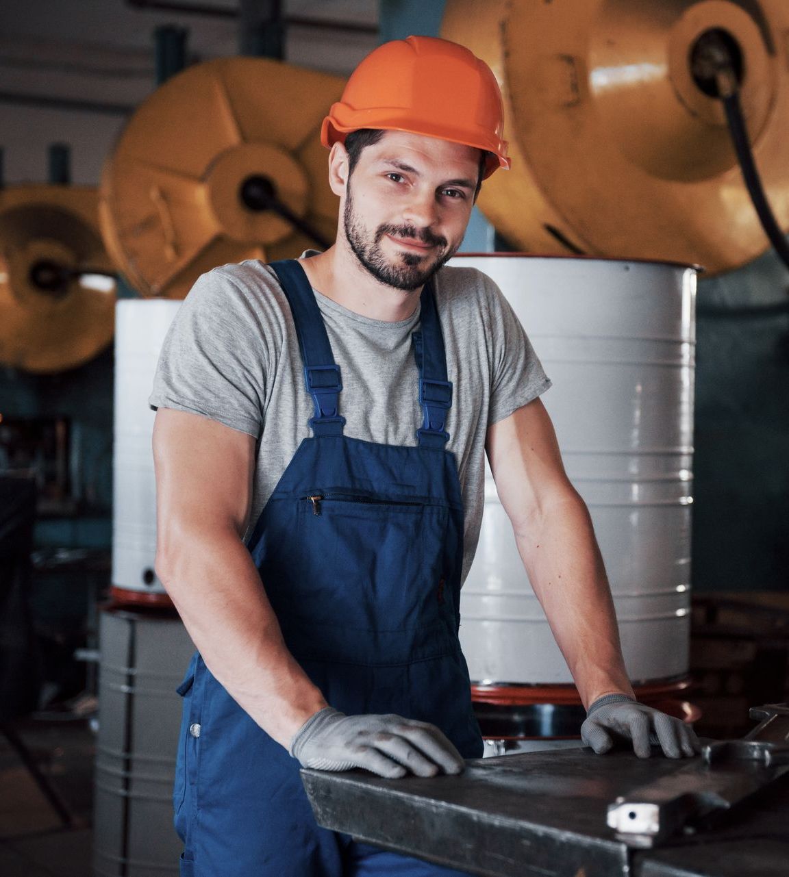 A man wearing a hard hat and overalls is standing in a factory.