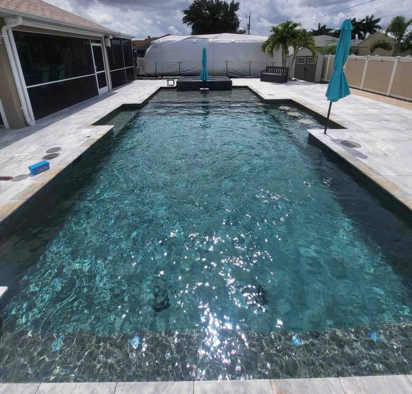Backyard swimming pool with turquoise water, white stone deck, and teal umbrellas under a cloudy sky
