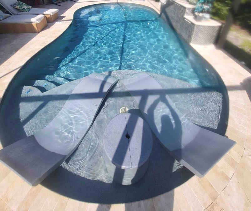 Round patio fire pit table beside a backyard swimming pool, with two black lounge chairs nearby.