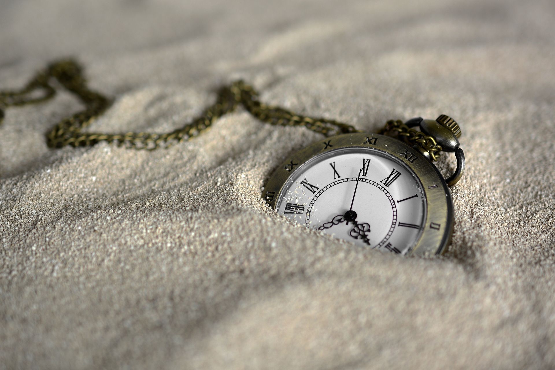 Pocket watch half-buried in sand on a beach, with chain trailing across the surface