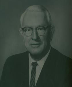 Black-and-white portrait of a man in glasses, suit, and tie against a plain background