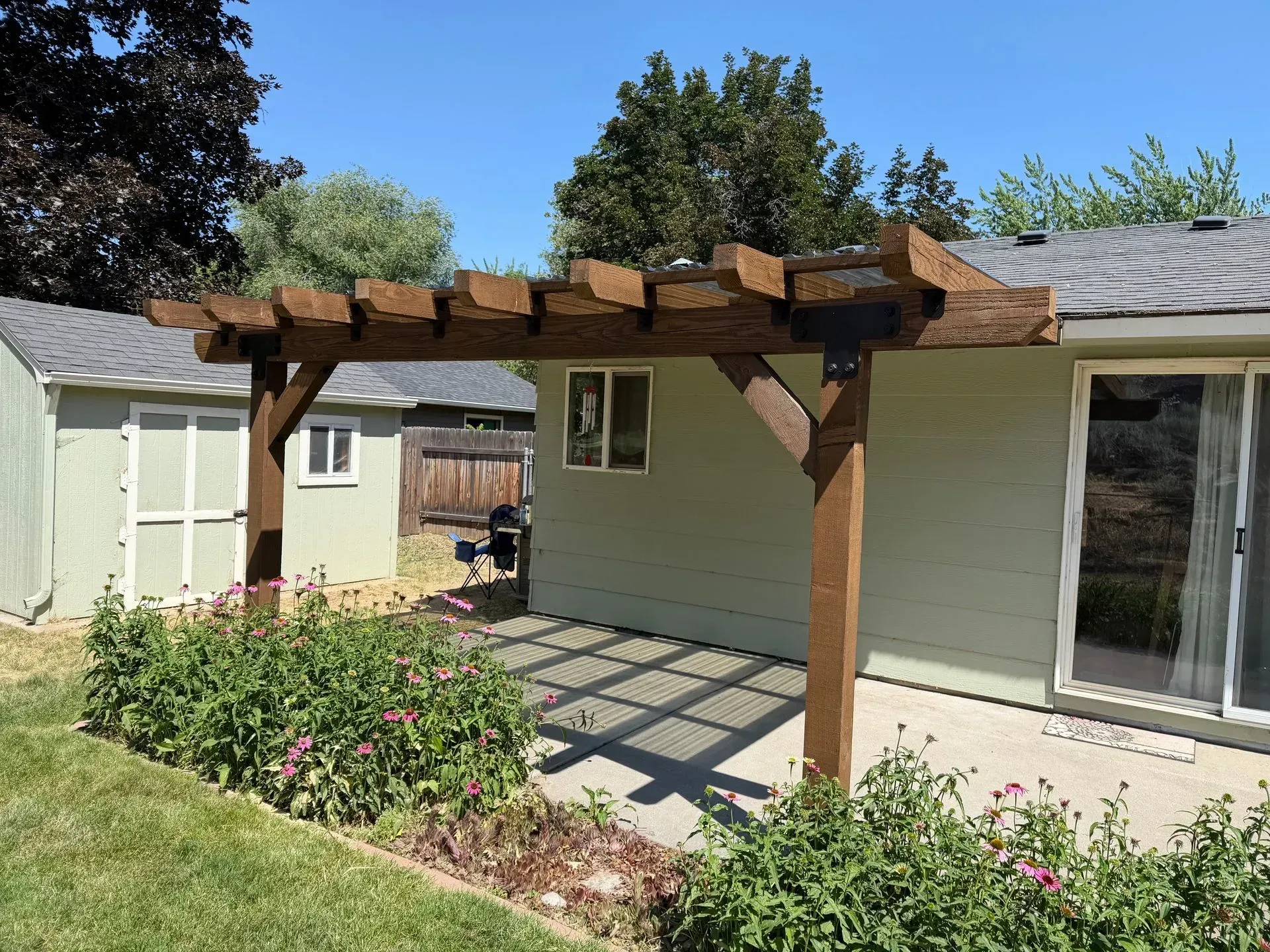 Backyard patio with a wooden pergola attached to a light-colored house, beside sliding glass doors.