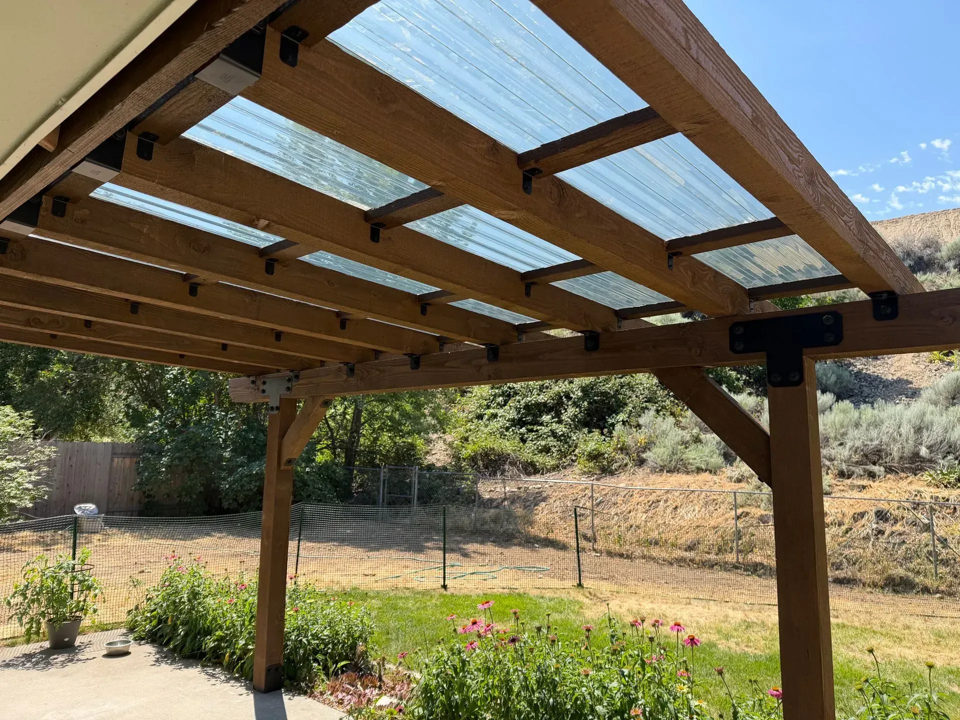 Wooden pergola with translucent roof panels over a garden, set against a sunny hillside.