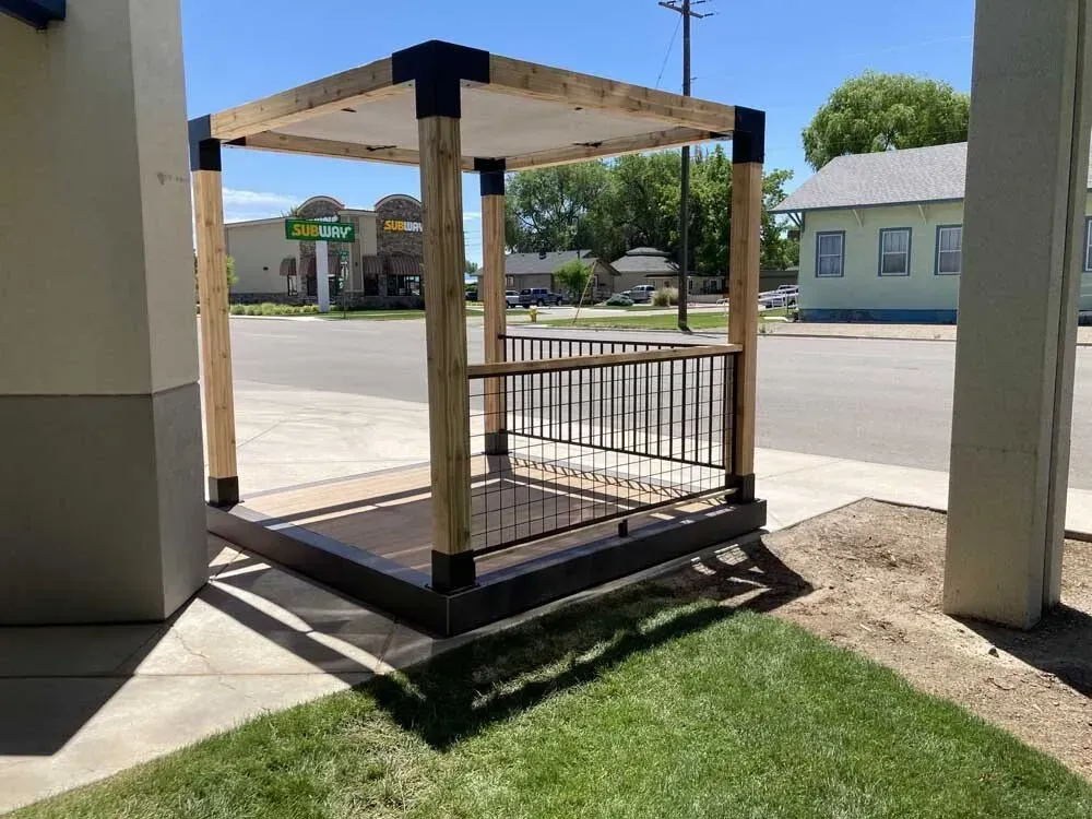 Covered outdoor bench with railings on a small concrete and grass platform beside a building entrance.