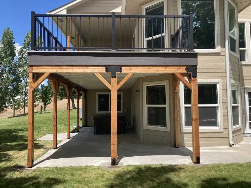 Covered patio under a second-story deck beside a beige house, with wood posts and black railings