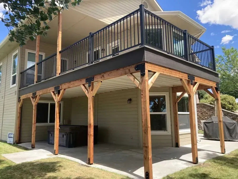 Raised backyard deck with black railing over a covered patio and wooden support posts, beside a house.