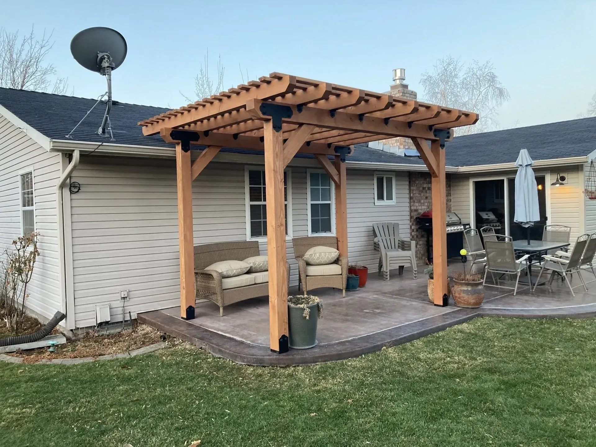 Wooden pergola over a backyard patio with chairs beside a brick house and green lawn