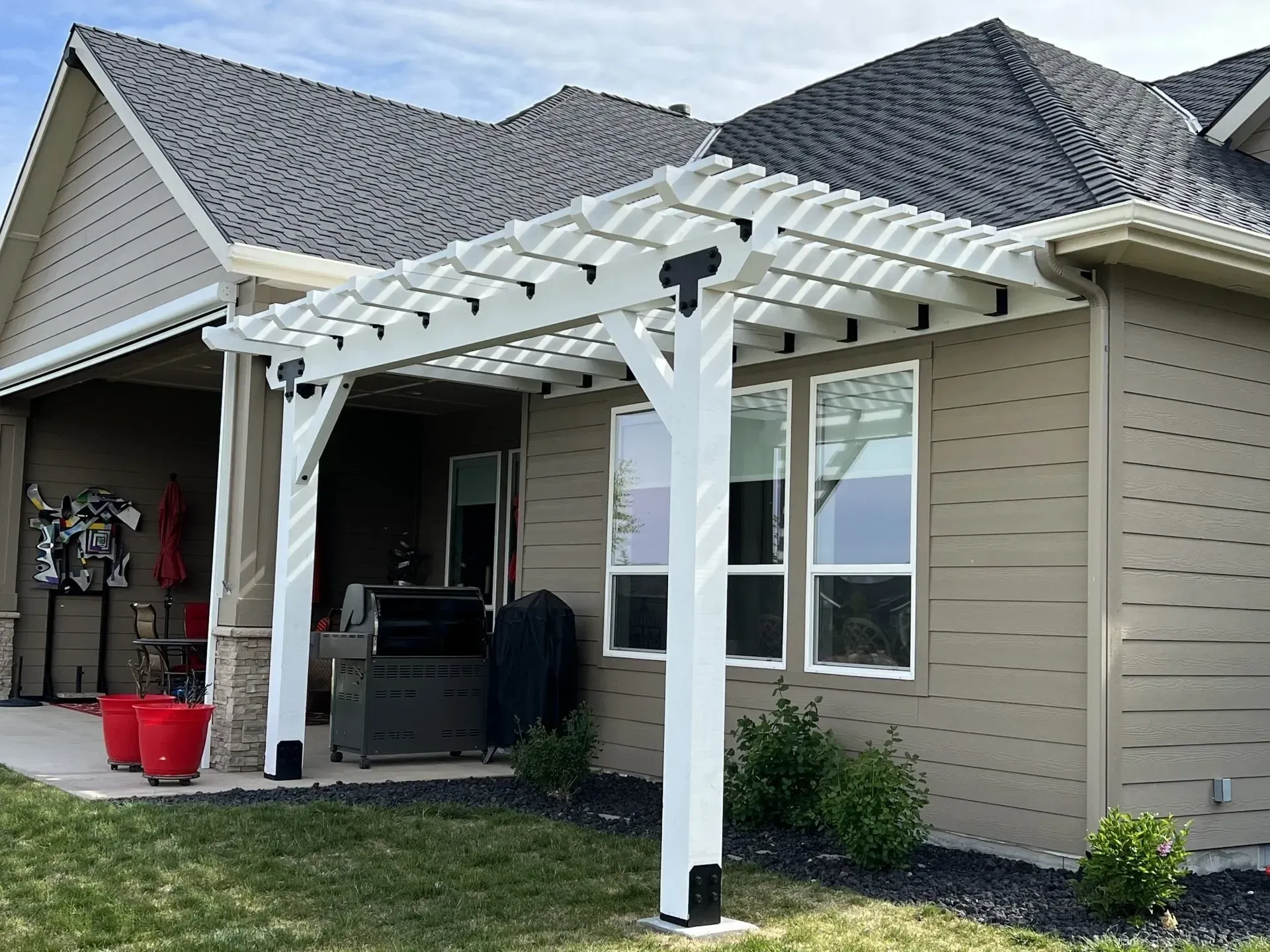 White pergola over a patio beside a tan house, with black grill and red buckets in front yard