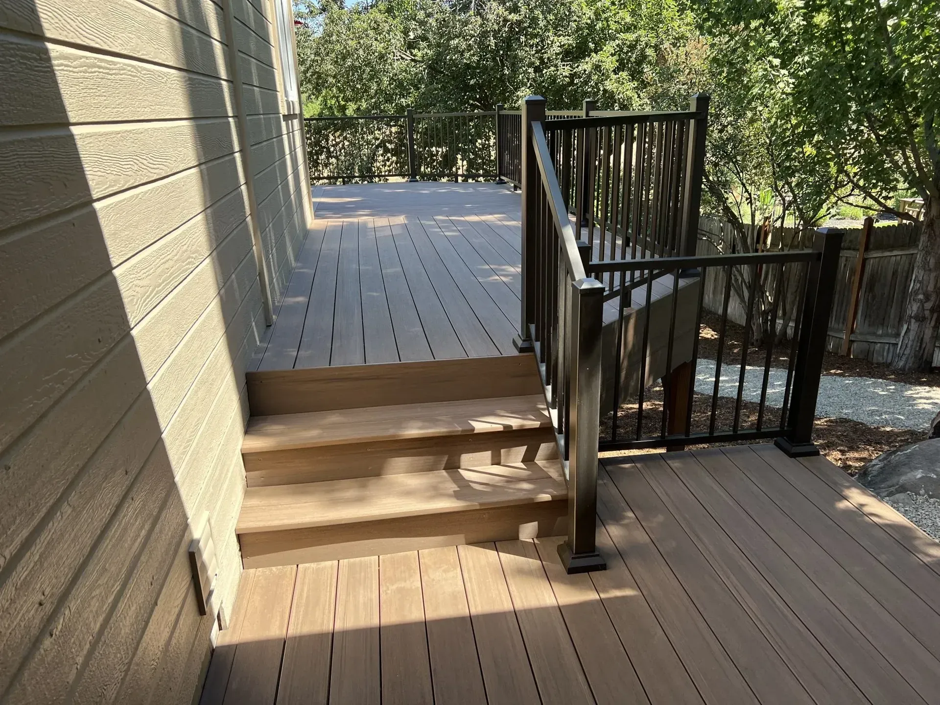 Wooden deck with steps and black railing beside a house, overlooking trees and a lower patio