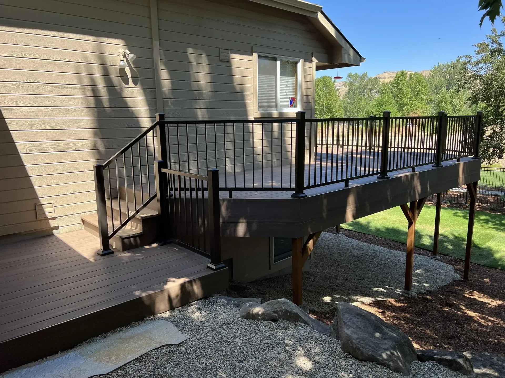 Raised wooden deck and ramp with black railings attached to a beige house, beside rocks and a yard.
