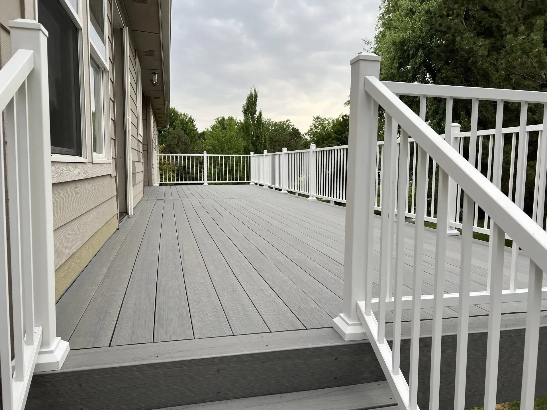 Wooden deck with white railing beside a house, overlooking trees under a cloudy sky
