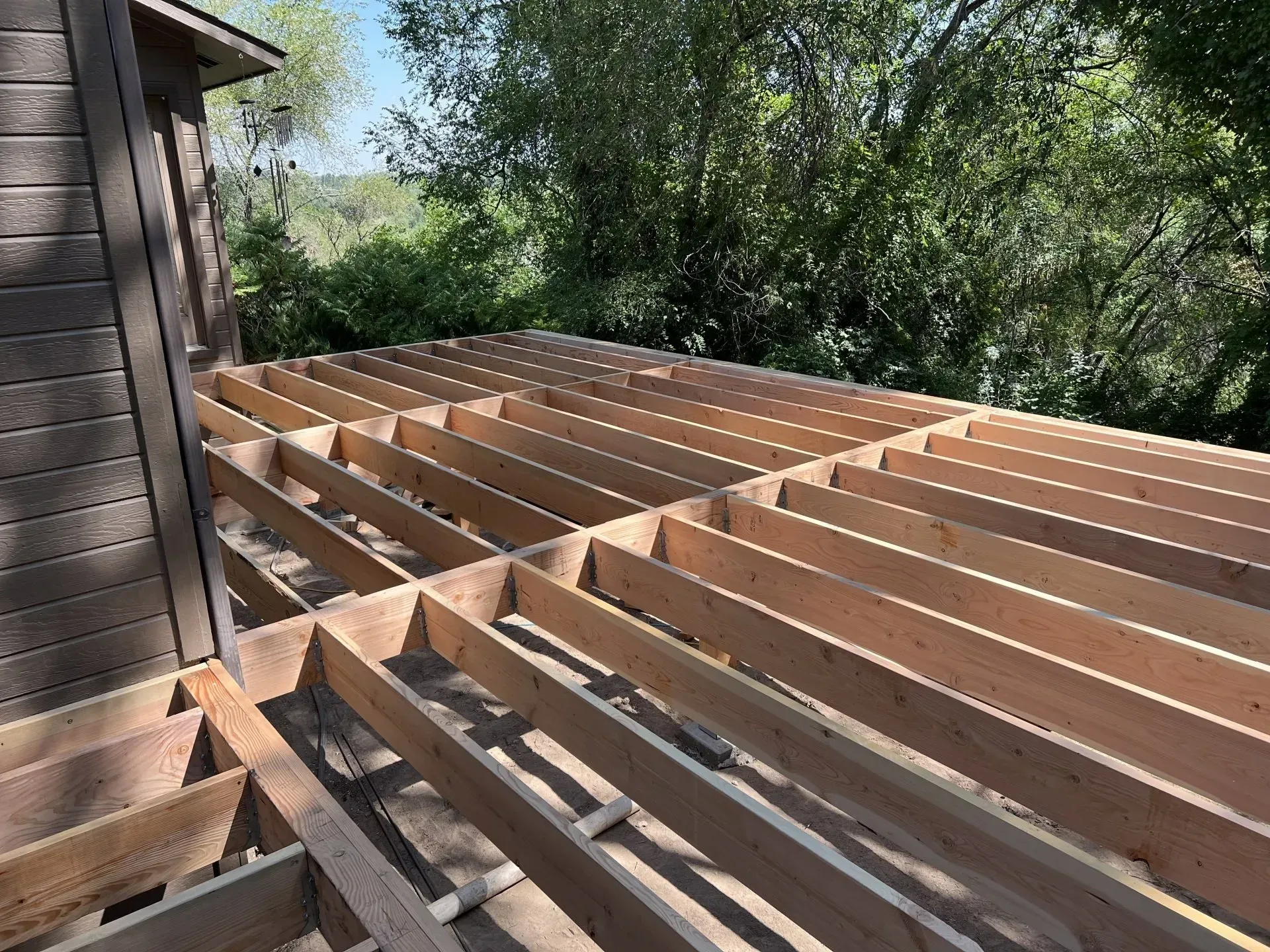 Wooden deck framing under construction beside a house, with exposed beams in a leafy backyard.