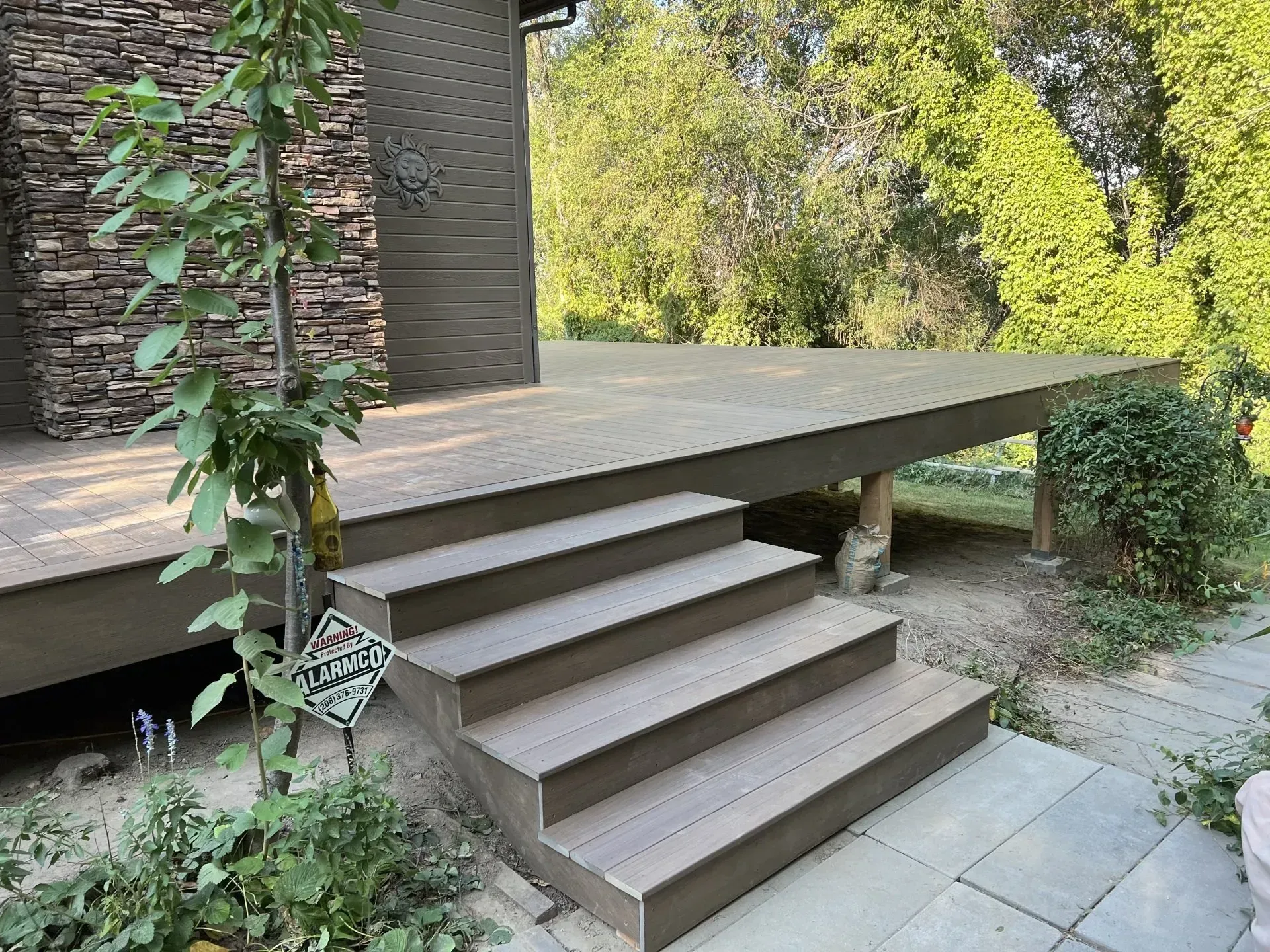 Wooden deck with steps leading to a raised patio beside a brick wall and garden greenery