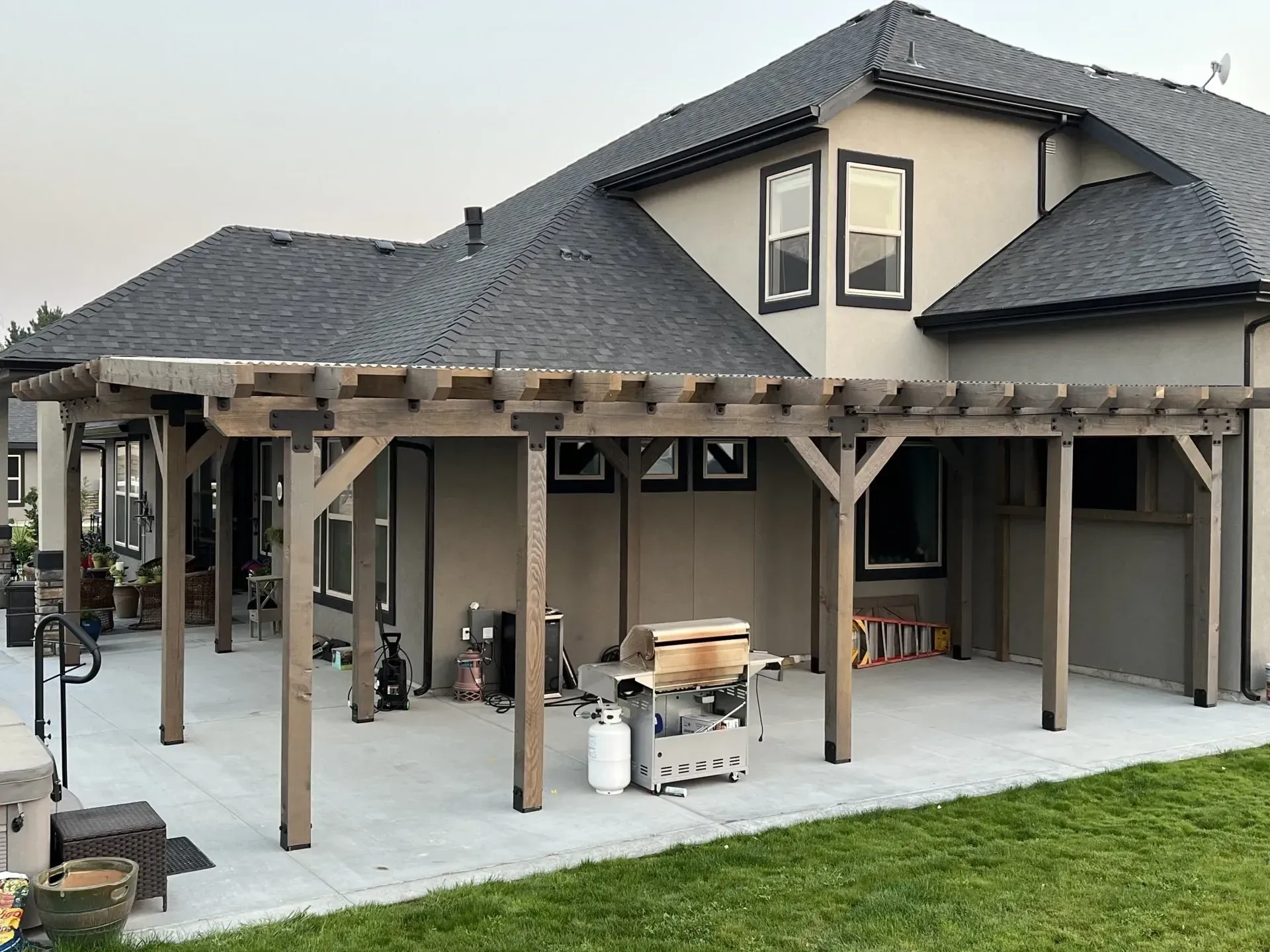 Covered patio with wooden pergola and stone barbecue beside a gray house under a dark shingled roof