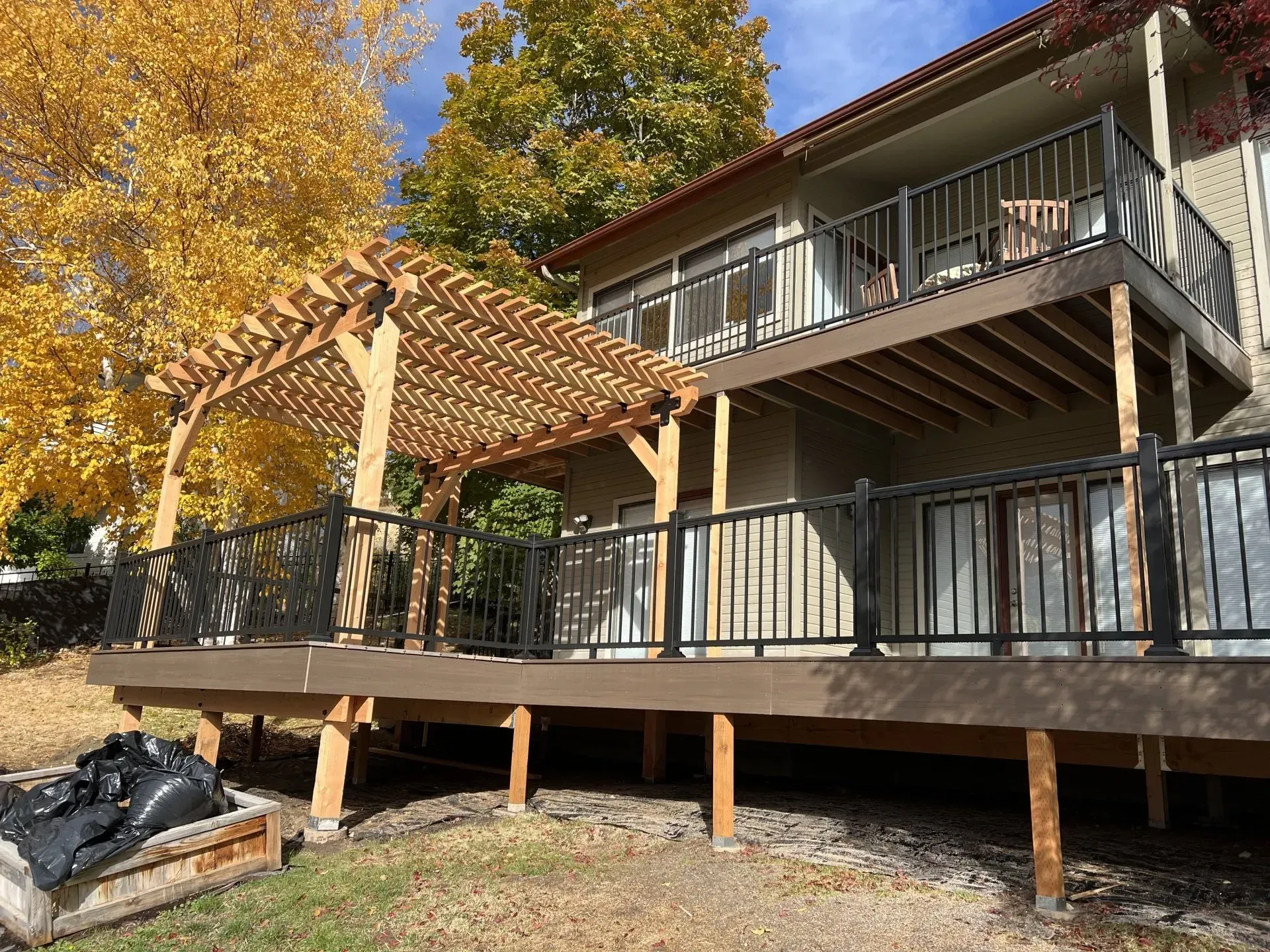 Covered wooden deck beside a house, with pergola, railing, and autumn trees in the yard.