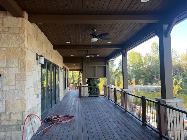 Covered stone patio with wooden deck, black railing, ceiling fan, and coiled hose beside glass doors