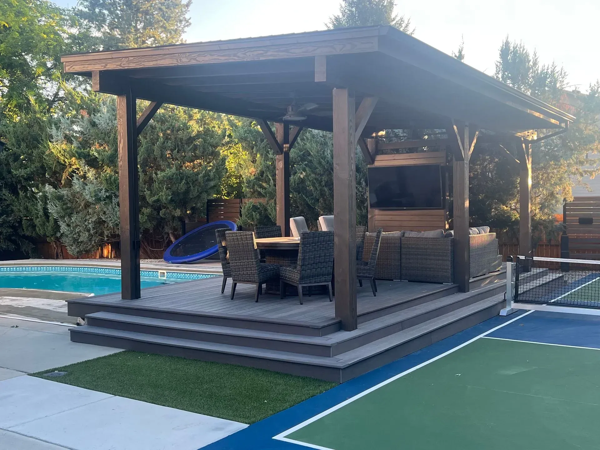 Backyard poolside patio with a wooden gazebo, outdoor seating, and steps beside a green court