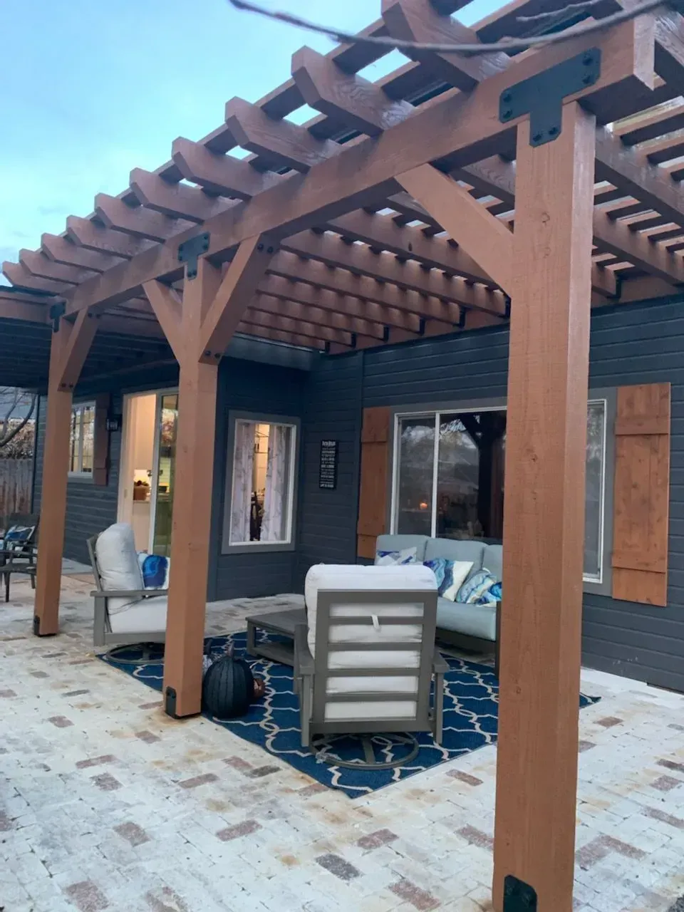 Covered patio with wooden pergola, gray seating, blue rug, and stone pavers beside a house.