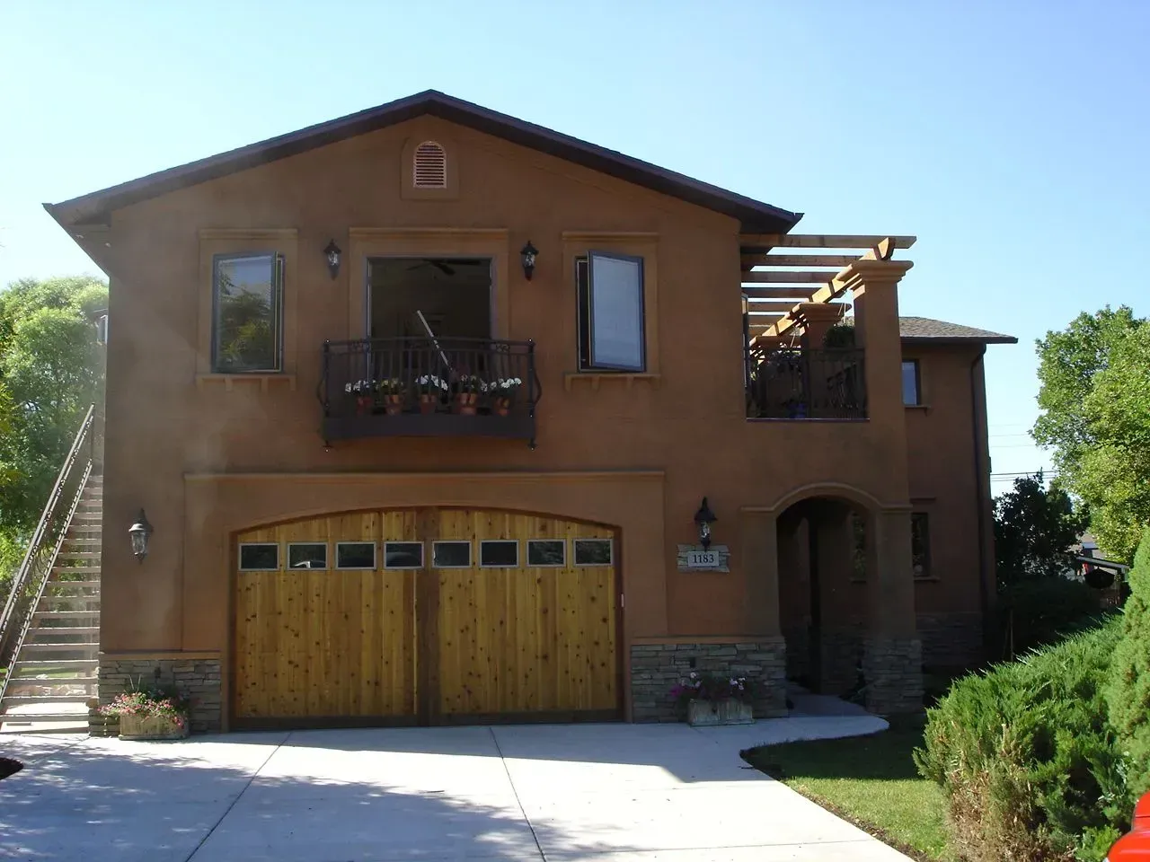 Two-story brick house with wooden garage doors, balcony, and side staircase on a sunny day