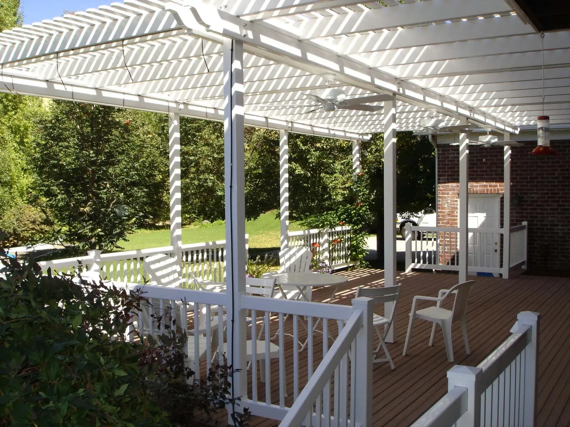 White pergola-covered deck with railing, white chairs, and a small table beside a brick house and greenery