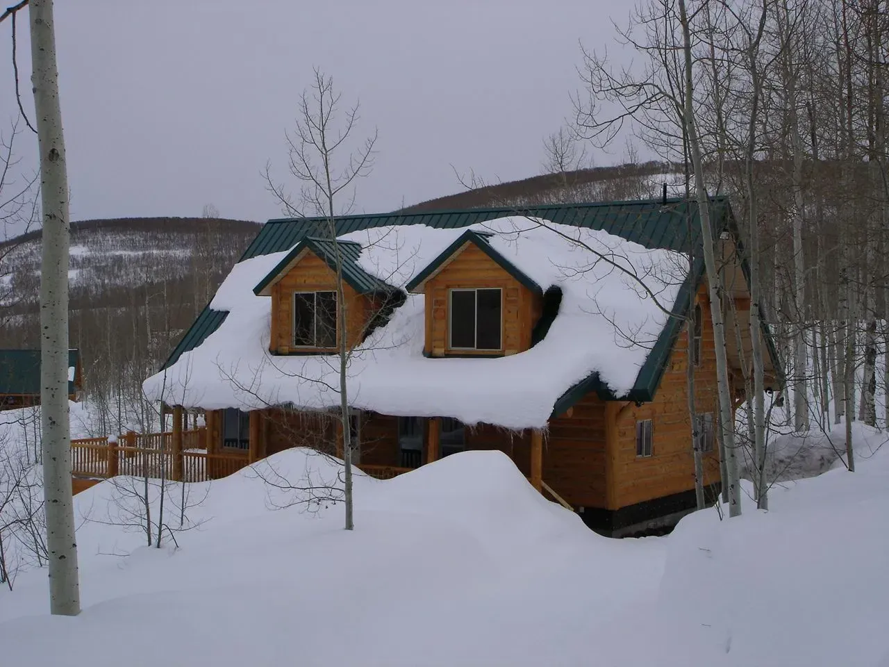 Snow-covered cabin in a wooded area, with a steep roof and dormer windows.