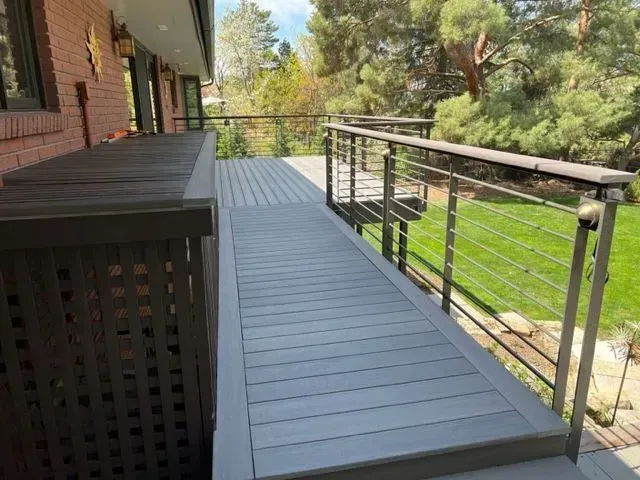 Wooden deck walkway beside a house, with black railing and trees in the background.