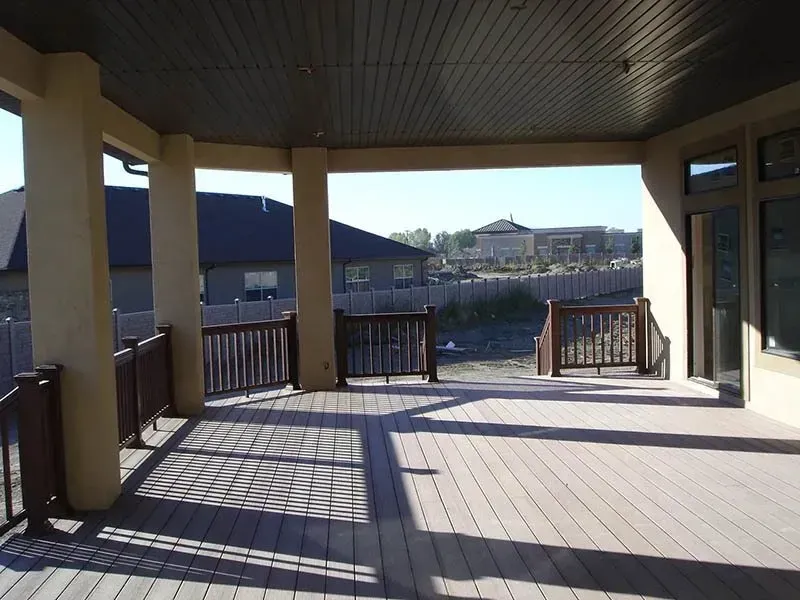 Covered wooden deck with railings and posts, overlooking water and buildings in daylight.