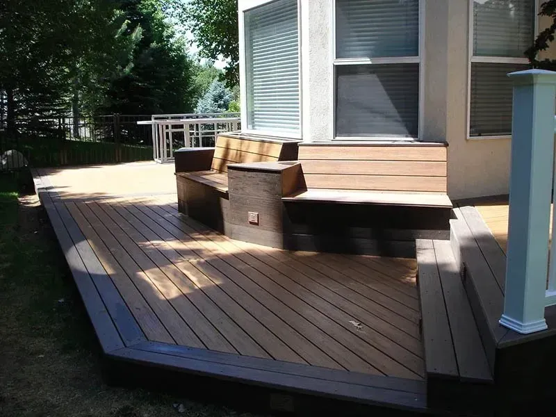 Wooden deck with built-in bench seating outside a house, bordered by trees and sunlight.