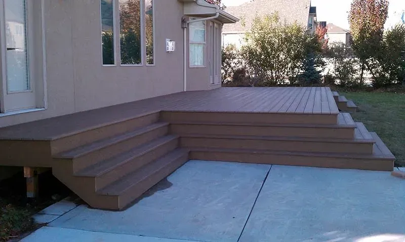 Wooden deck steps leading to a raised patio beside a house with a concrete walkway and lawn.