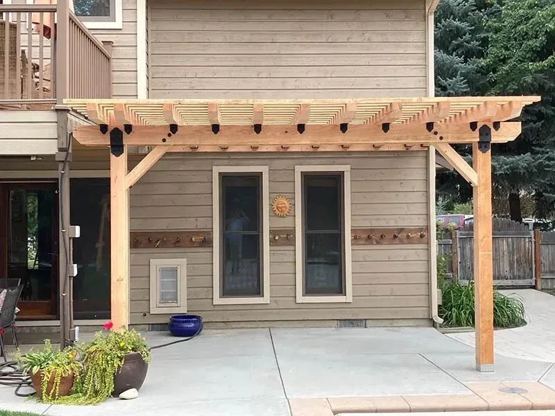Wooden pergola over a concrete patio beside a beige house with windows and potted plants