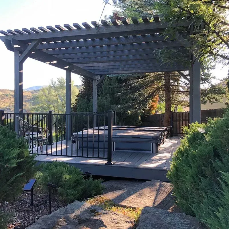 Backyard deck with a gray pergola, black railing, and cushioned seating surrounded by trees and shrubs