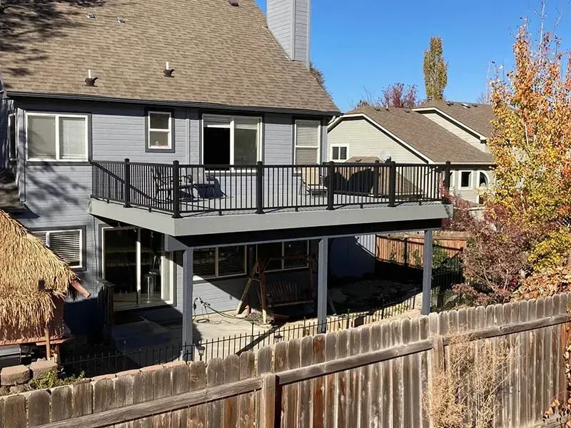 Gray house with a raised deck and black railing, viewed over a wooden fence in a residential yard
