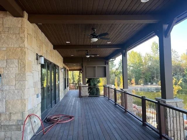 Covered wooden porch with stone wall, black railing, ceiling fan, and coiled red hose.
