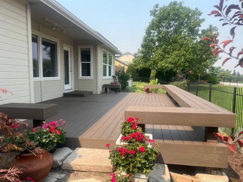 Spacious gray wooden deck beside a house, with built-in benches, potted flowers, and a green yard beyond.