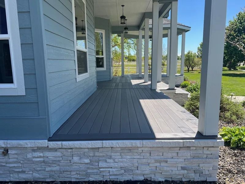 Covered front porch with gray siding, dark deck boards, white columns, and stone skirting overlooking a green yard