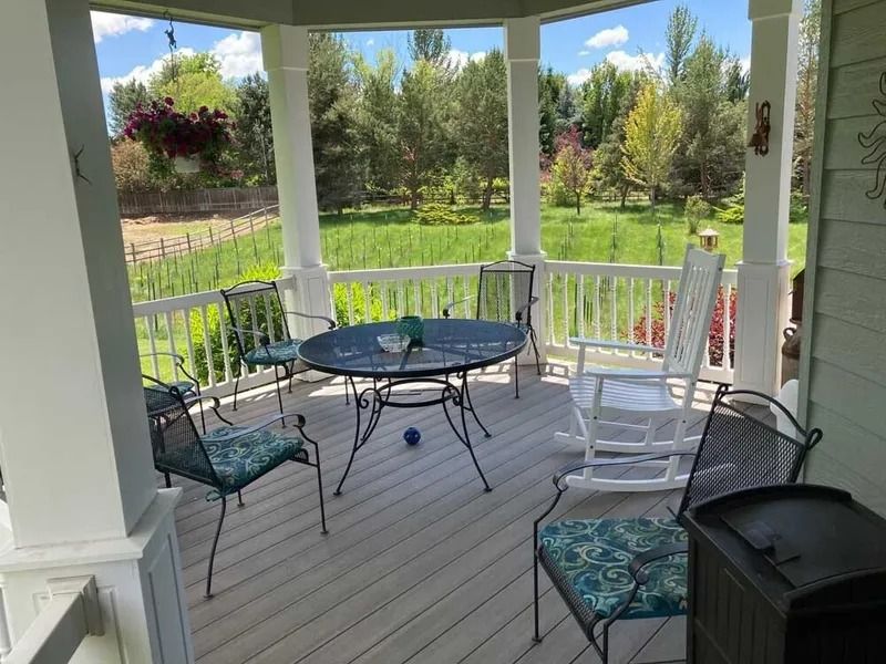 Covered porch with white rocking chairs, round table, hanging flowers, and a green garden view.