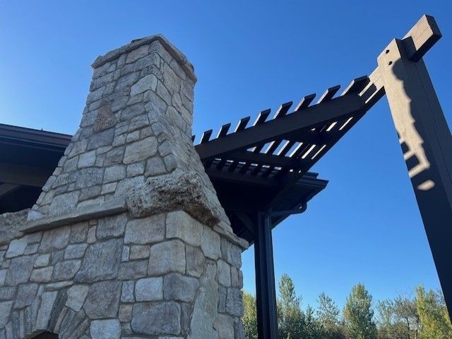 Stone chimney and dark wooden pergola against a bright blue sky.