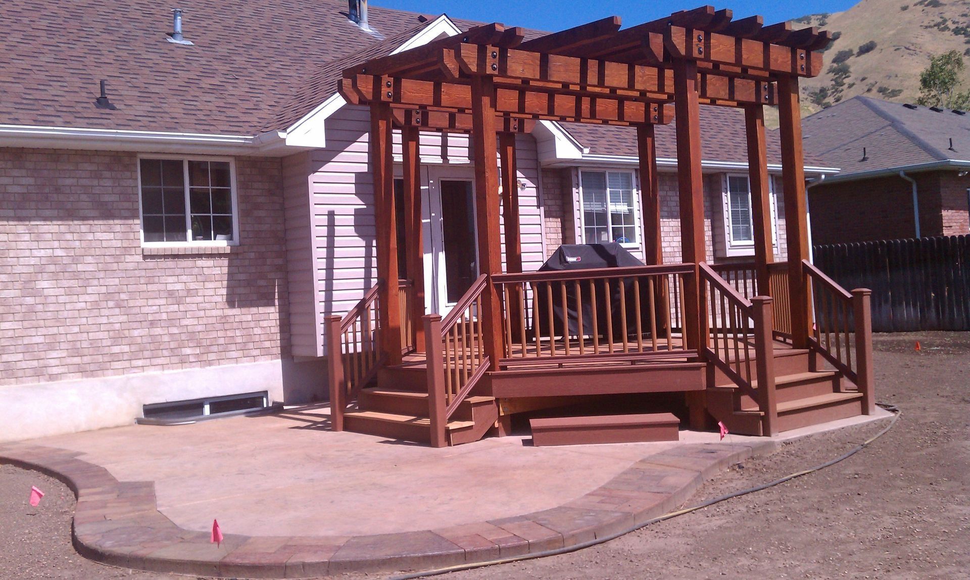 Wooden backyard gazebo on a concrete patio beside a brick house, with railings and a shingled roof.