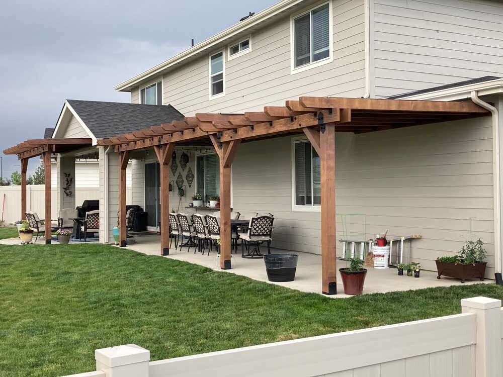 Backyard patio with wooden pergola, outdoor seating, and beige house siding beside green lawn.