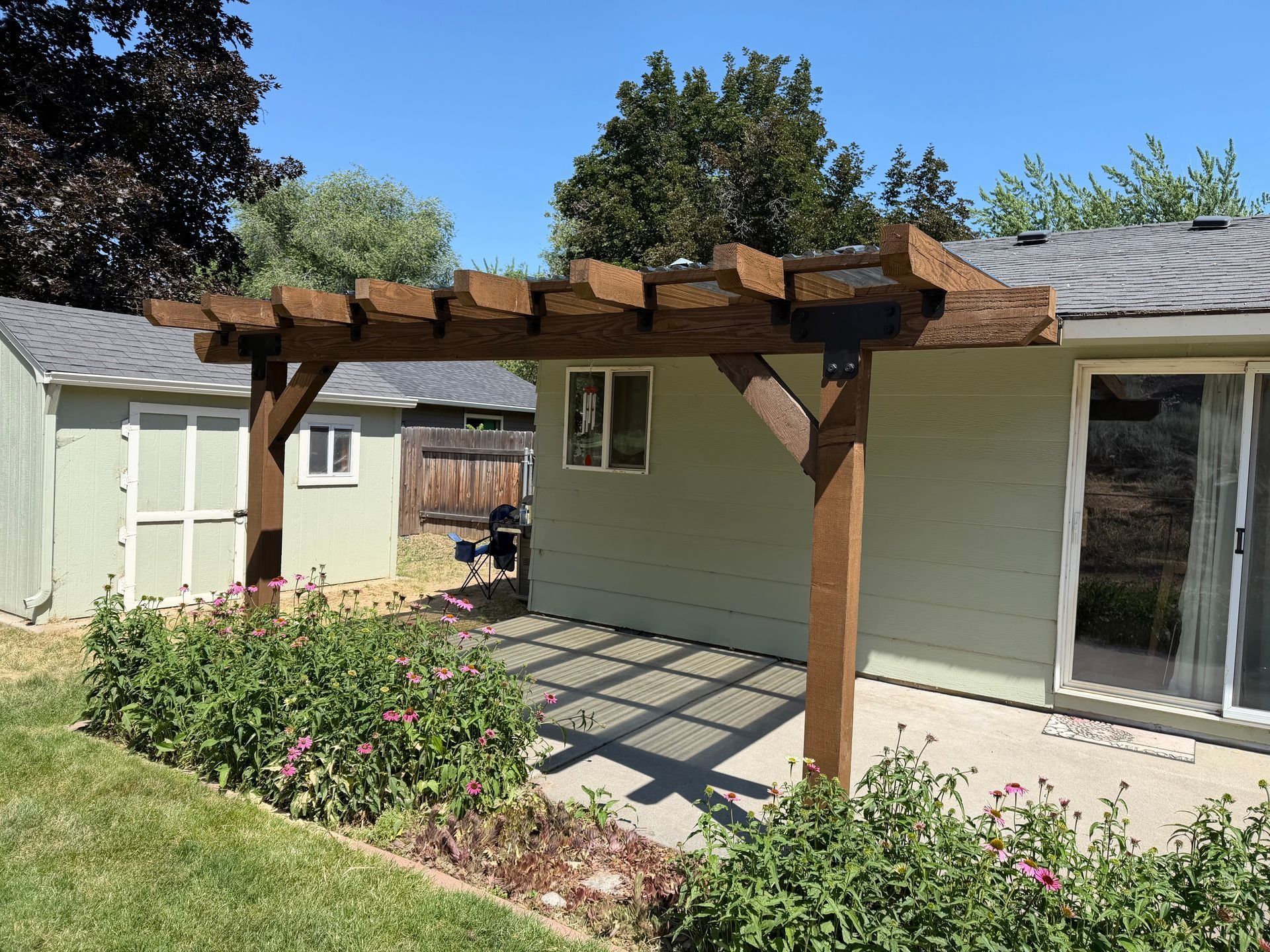 Backyard patio with wooden pergola attached to a light green house, beside a garden bed and sliding door