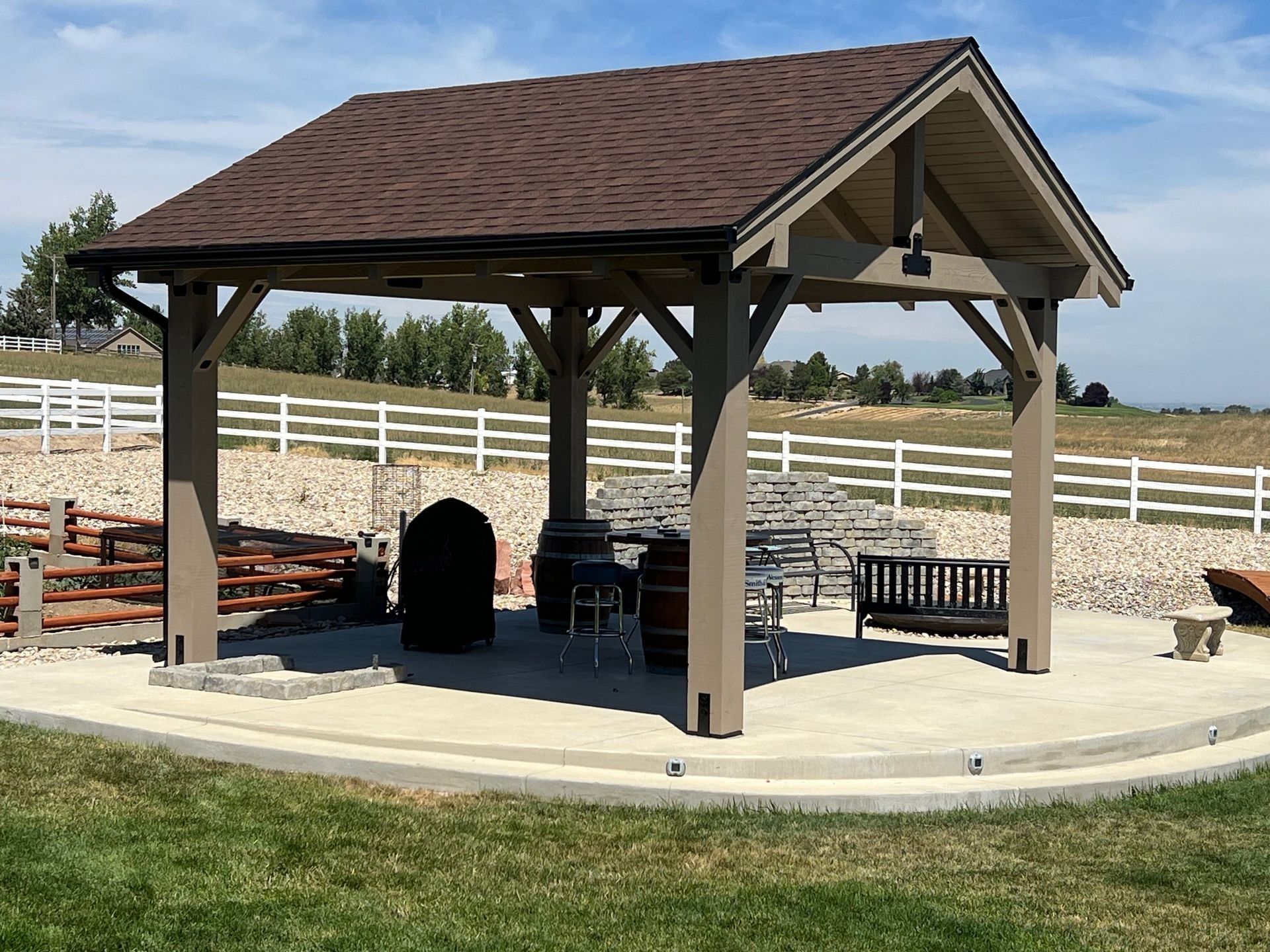 Covered pavilion with picnic tables and chairs on a concrete pad in a fenced grassy park.