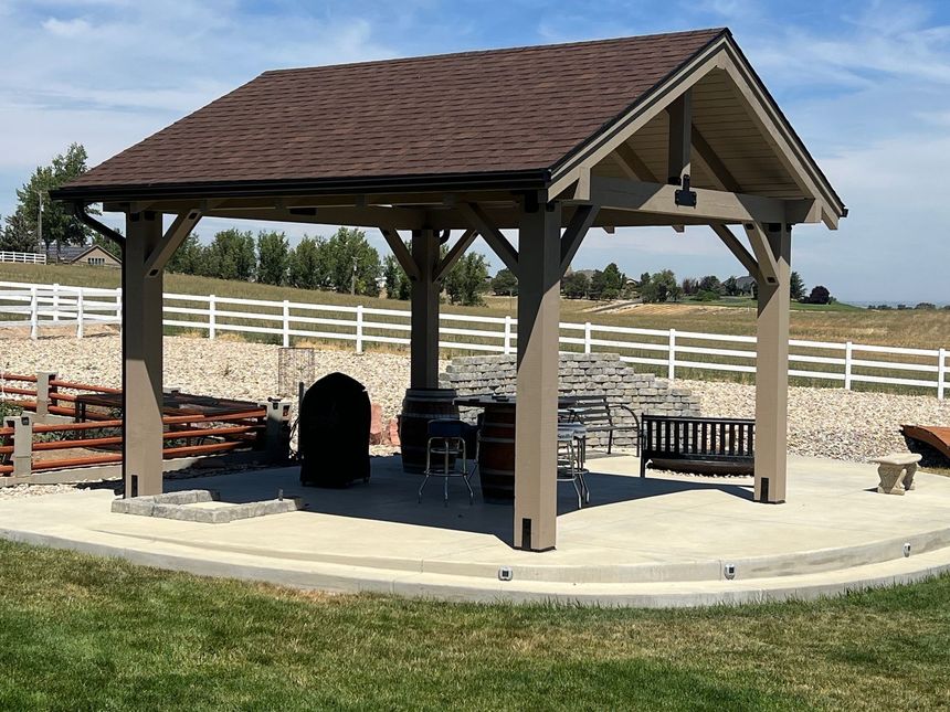 Outdoor pavilion with picnic tables, a grill, and benches on a concrete pad in a fenced field.