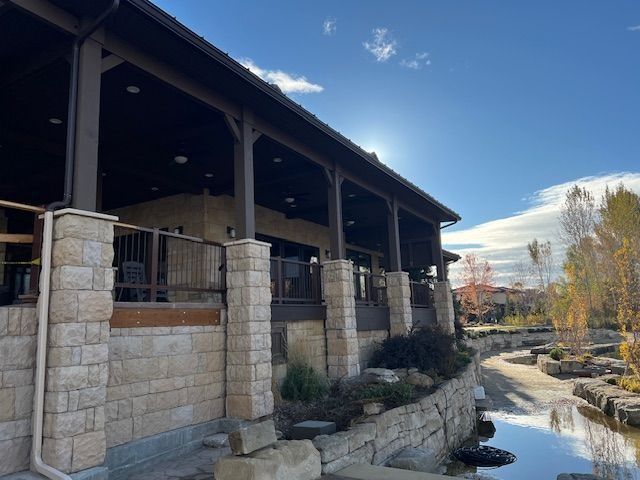 Stone building with wooden porch beside a narrow stream under a bright blue sky