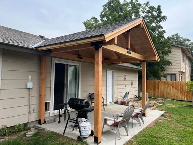 Covered backyard patio with wooden roof posts, grill, and chairs beside a house.