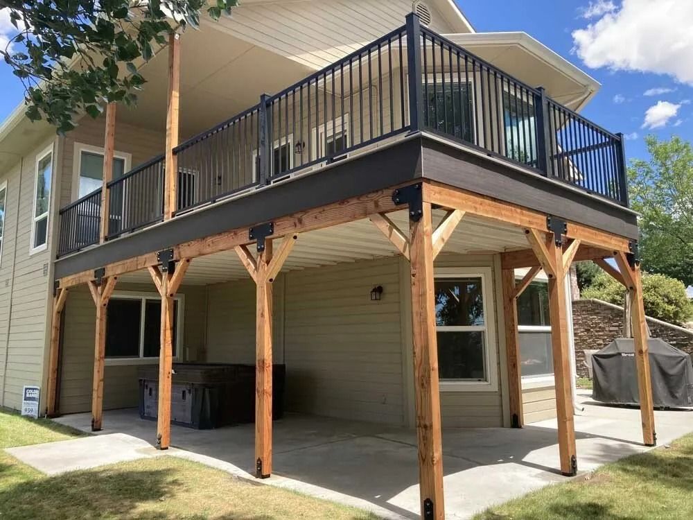 Raised beige house with black balcony and wooden posts over a covered patio and driveway