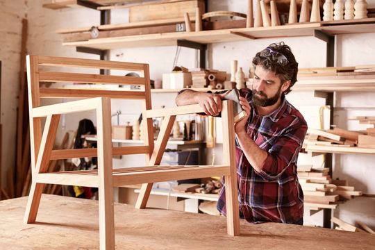 Man in plaid shirt working with a power drill on a wooden chair in a woodworking workshop