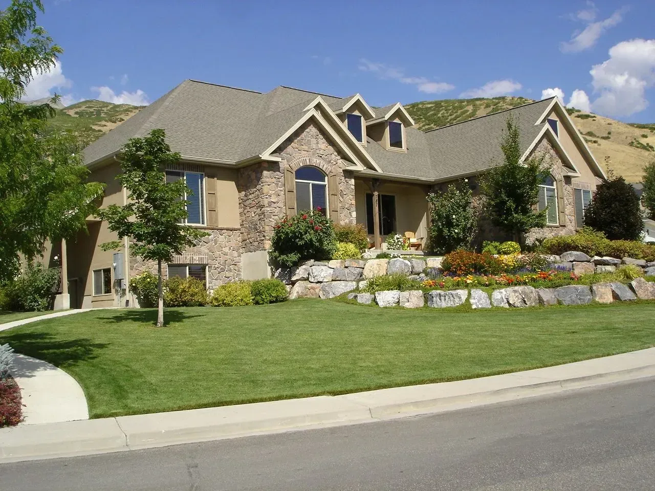 Large stone house with a manicured lawn, flower beds, and a curved driveway under a blue sky