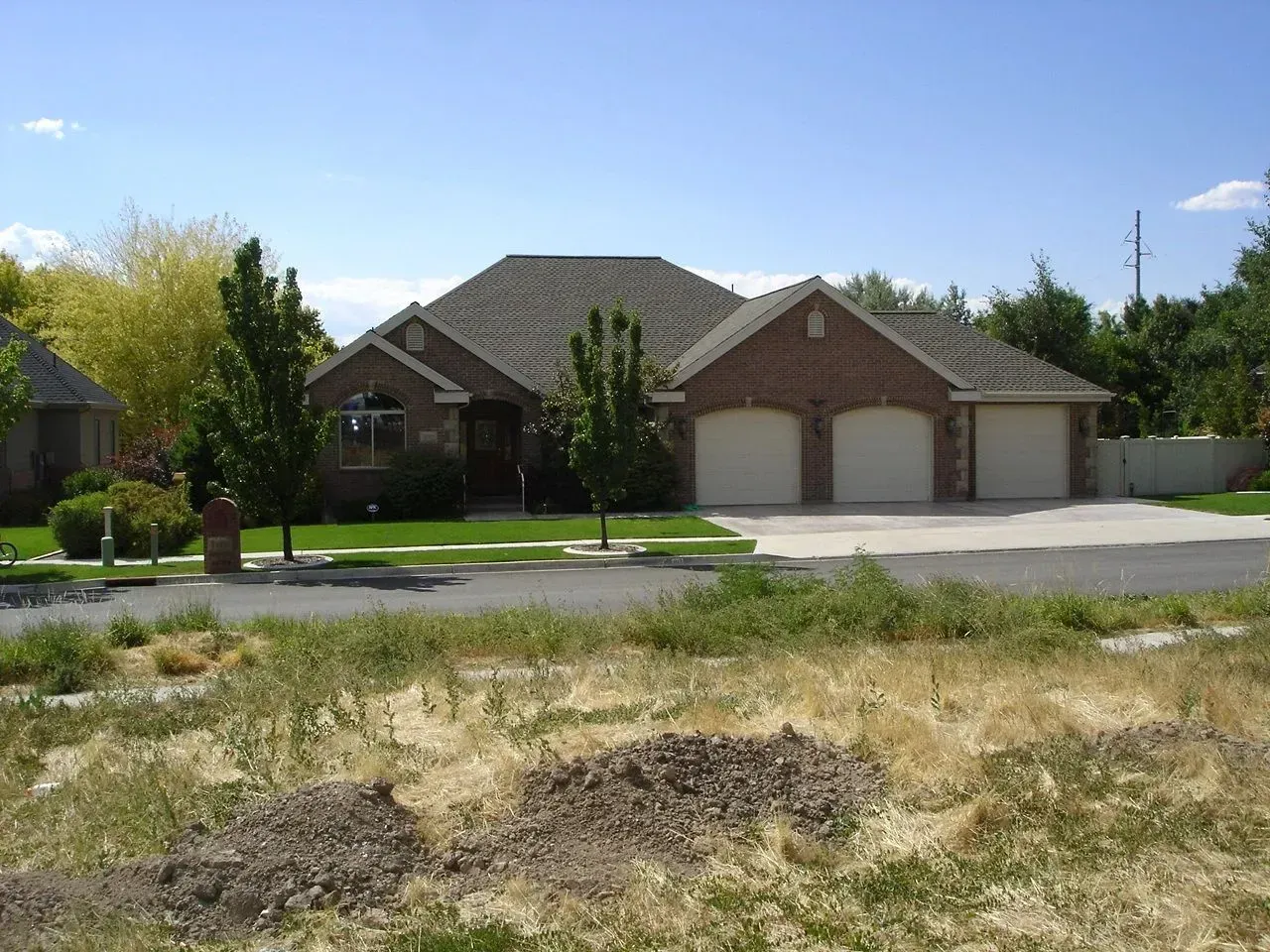 Suburban brick house with a two-car garage beside a grassy, undeveloped lot under a blue sky
