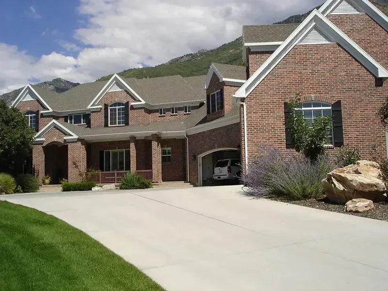 Suburban brick house with a large driveway, green lawn, and mountain backdrop under a partly cloudy sky